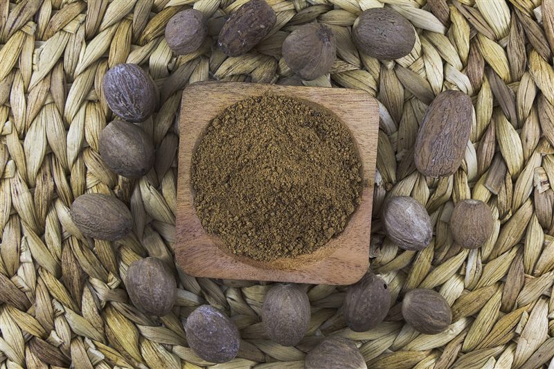 Whole brown nutmegs arranged around a small wooden dish of ground nutmeg on a woven mat